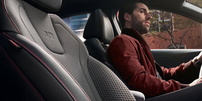 Hombre con barba y chaqueta marrón conduciendo un coche con asientos de cuero negro y gris con costuras rojas.