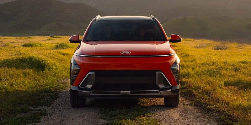 Vista frontal de un Hyundai Exter rojo, estacionado en un camino de tierra en un campo de hierba con colinas al fondo.