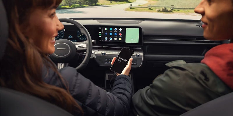 Dos personas en un coche, una mujer al volante y un hombre a su lado, mirando un teléfono inteligente con "Android Auto" en la pantalla.