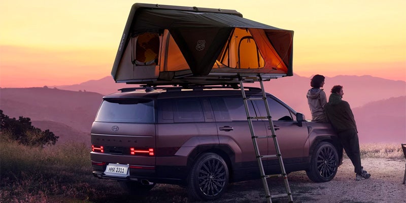Un SUV Hyundai Santa Fe con tienda de techo y escalera, estacionado con dos personas en la puerta trasera contemplando una puesta de sol sobre las montañas.