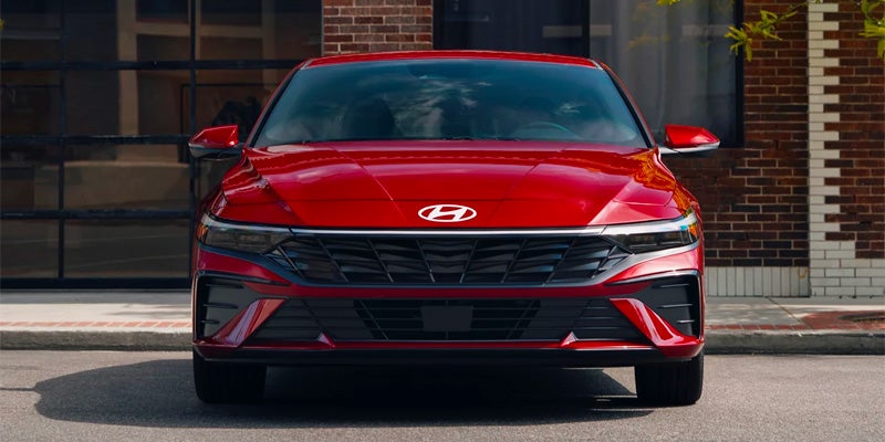 Vista frontal de un coche Hyundai rojo aparcado en la calle, con un edificio de ladrillos al fondo.