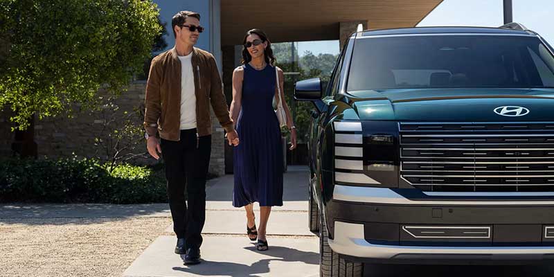 Una pareja, tomada de la mano, camina junto a un SUV Hyundai Palisade de color verde oscuro en dirección a una casa moderna.